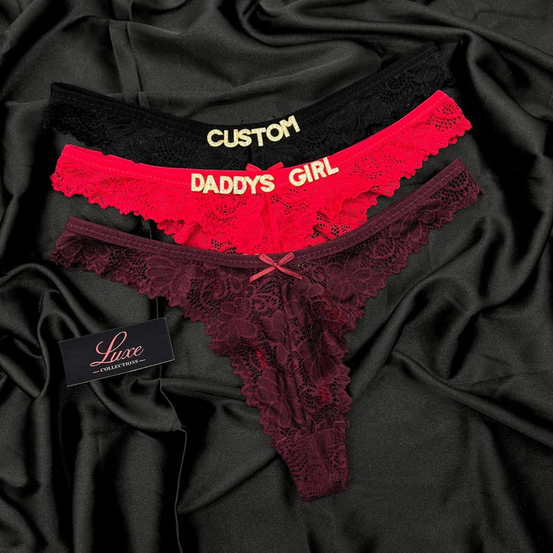 Pode incluir: Tr&ecirc;s tangas de renda nas cores preta, vermelha e bord&ocirc; escuro. O par preto apresenta o texto dourado CUSTOM, o par vermelho apresenta o texto dourado DADDYS GIRL e o par bord&ocirc; tem um pequeno la&ccedil;o. Uma etiqueta preta exibe Luxe COLLECTIONS.