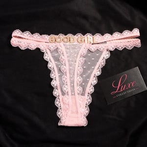 K&ouml;nnte beinhalten: Ein hellrosa Tanga aus transparentem Netzstoff mit Schweizer Punkten, verziert mit gewellter Spitzenborte und einem mit Strass besetzten Metallanh&auml;nger, der die Worte GOOD GIRL am Bund bildet. Eine schwarze Karte mit dem Text Luxe COLLECTIONS liegt bei.