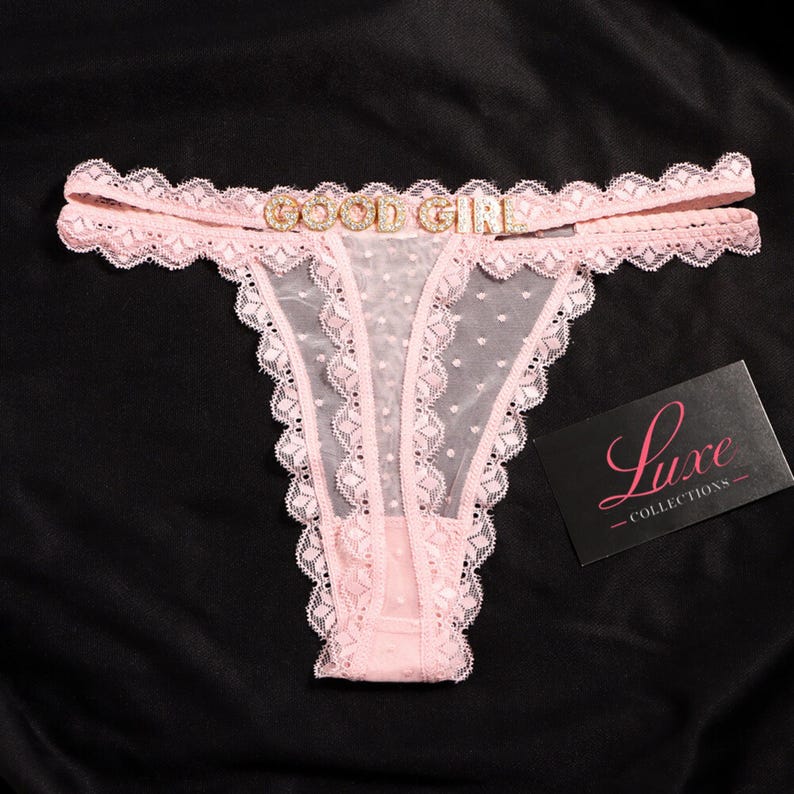 K&ouml;nnte beinhalten: Ein hellrosa Tanga aus transparentem Netzstoff mit Schweizer Punkten, verziert mit gewellter Spitzenborte und einem mit Strass besetzten Metallanh&auml;nger, der die Worte GOOD GIRL am Bund bildet. Eine schwarze Karte mit dem Text Luxe COLLECTIONS liegt bei.