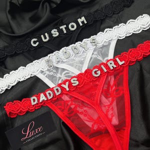 Pode incluir: Três tangas de renda nas cores preta, branca e vermelha com letras de strass. O par preto exibe a palavra CUSTOM, o par branco exibe DADDY'S e o par vermelho exibe DADDYS GIRL. Cada peça possui um cós de renda com acabamento festonado.