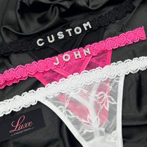 May include: Three thong underwear pieces in black, hot pink, and white, each featuring a heart-patterned lace waistband. The black thong displays the word CUSTOM in rhinestones, the pink thong displays JOHN in rhinestones, and a Luxe Collections tag.