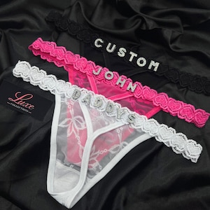 Puede incluir: Tres tangas en blanco, rosa y negro, cada uno con una cintura de encaje con letras de pedrer&iacute;a. El par blanco deletrea DADDYS, el rosa JOHN y el negro CUSTOM. Se incluye una etiqueta negra con el texto Luxe Collections en la imagen.