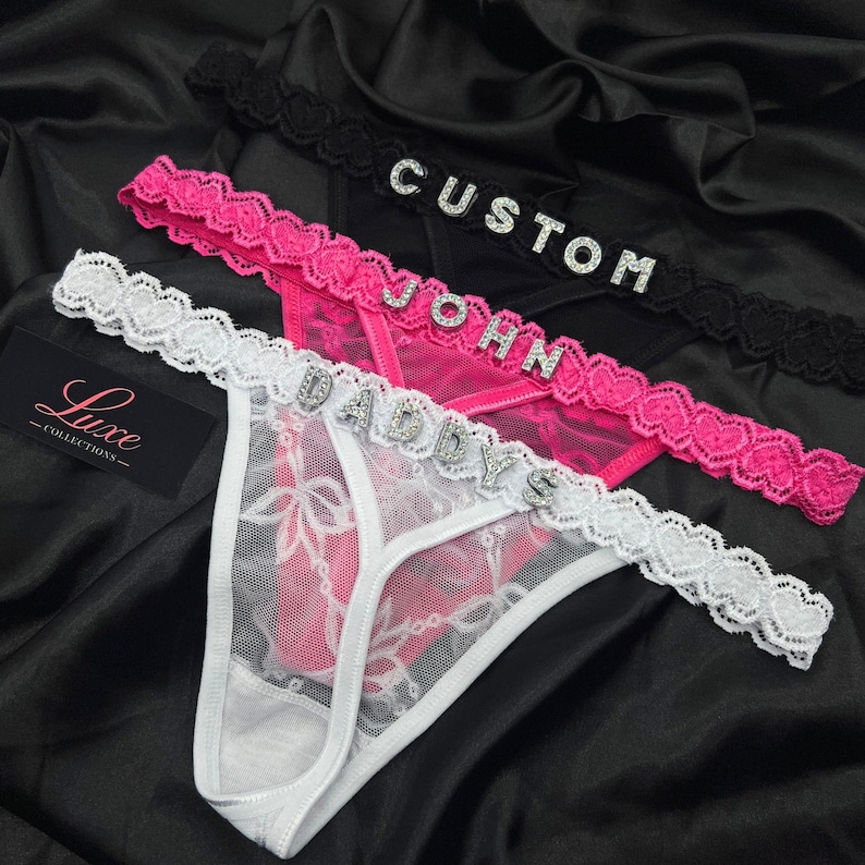 Puede incluir: Tres tangas en blanco, rosa y negro, cada uno con una cintura de encaje con letras de pedrer&iacute;a. El par blanco deletrea DADDYS, el rosa JOHN y el negro CUSTOM. Se incluye una etiqueta negra con el texto Luxe Collections en la imagen.