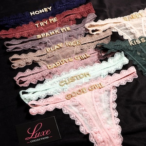 K&ouml;nnte beinhalten: Eine Kollektion von Spitzen-Tangas in verschiedenen Farben wie Marineblau, Burgunderrot, Mauve, Beige, Rosa, Mint und Dunkelgr&uuml;n. Jeder Tanga ist mit goldfarbenen Strassbuchstaben verziert, die W&ouml;rter wie HONEY, TRY ME, SPANK ME, PLAY NICE, DADDYS GIRL, CUSTOM, GOOD GIRL, DADDY und KISS bilden. Eine schwarze Karte mit dem Text Luxe COLLECTIONS ist zu sehen.