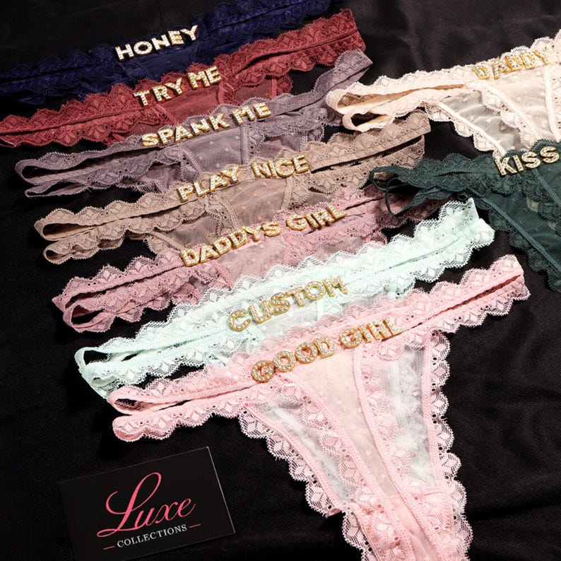 K&ouml;nnte beinhalten: Eine Kollektion von Spitzen-Tangas in verschiedenen Farben wie Marineblau, Burgunderrot, Mauve, Beige, Rosa, Mint und Dunkelgr&uuml;n. Jeder Tanga ist mit goldfarbenen Strassbuchstaben verziert, die W&ouml;rter wie HONEY, TRY ME, SPANK ME, PLAY NICE, DADDYS GIRL, CUSTOM, GOOD GIRL, DADDY und KISS bilden. Eine schwarze Karte mit dem Text Luxe COLLECTIONS ist zu sehen.