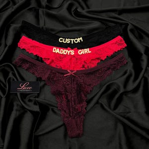 Pode incluir: Tr&ecirc;s tangas de renda nas cores preta, vermelha e bord&ocirc; escuro. O par preto apresenta o texto dourado CUSTOM, o par vermelho o texto dourado DADDYS GIRL e o par bord&ocirc; um pequeno la&ccedil;o central. Uma etiqueta preta exibe Luxe COLLECTIONS.