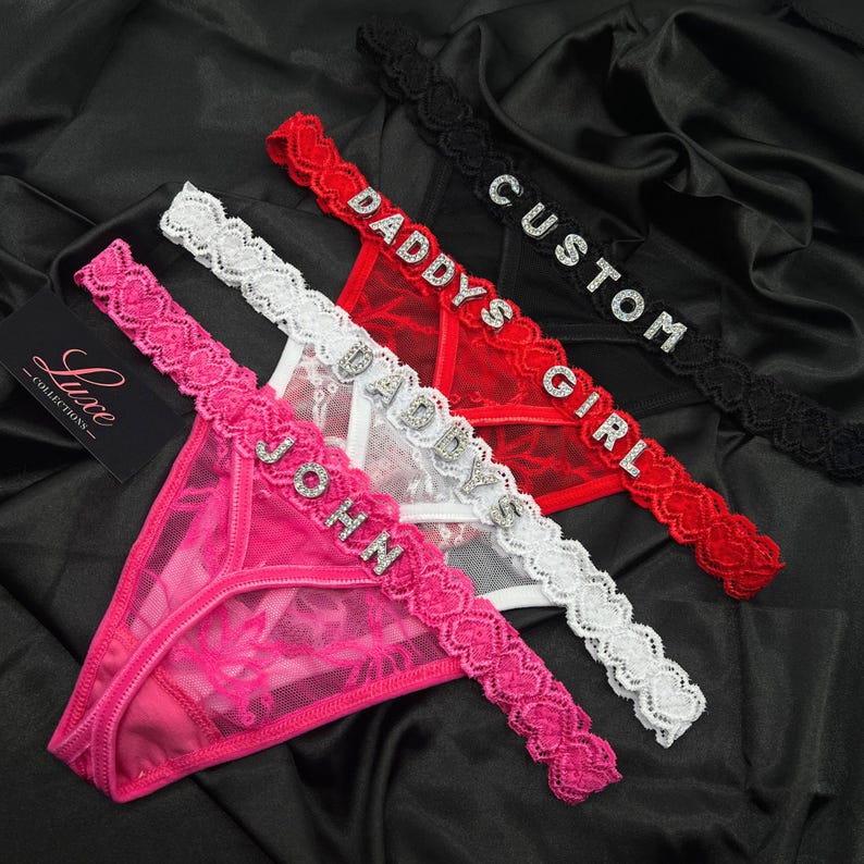 Puede incluir: Tres tangas en rosa, blanco y rojo, cada uno con una cintura de encaje festoneado con letras de diamantes de imitaci&oacute;n. El tanga rosa dice JOHN, el blanco dice DADDY y el rojo dice DADDYS GIRL. Una etiqueta negra tiene la palabra Luxe.