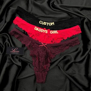 Pode incluir: Tr&ecirc;s tangas de renda nas cores preta, vermelha e bord&ocirc; escuro. A tanga preta apresenta o texto dourado CUSTOM, a vermelha o texto dourado DADDYS GIRL e a bord&ocirc; um pequeno la&ccedil;o central. Uma etiqueta preta exibe o texto Luxe COLLECTIONS.