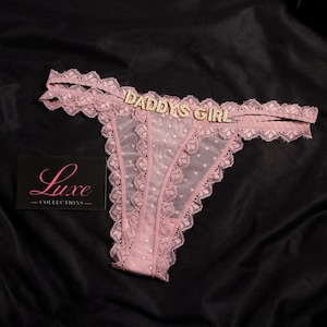 K&ouml;nnte beinhalten: Altrosa Tanga aus transparentem Netzstoff mit Punkten, Spitzenbesatz und goldfarbenen Strassbuchstaben, die DADDYS GIRL auf dem vorderen Bund bilden. Eine schwarze Karte mit dem Text Luxe Collections ist ebenfalls abgebildet.