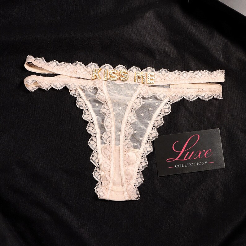 K&ouml;nnte beinhalten: Ein hellbeiger Spitzen-String mit einem transparenten Mesh-Einsatz mit Punkten und einem doppelten Bund. Die Vorderseite ist mit goldfarbenen Metallbuchstaben verziert, die KISS ME buchstabieren. Eine schwarze Karte mit dem Text Luxe COLLECTIONS liegt bei.