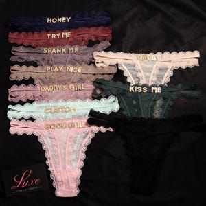 K&ouml;nnte beinhalten: Eine Kollektion von Spitzen-Tangas in verschiedenen Farben wie Marineblau, Bordeaux, Mauve, Beige, Dunkelgr&uuml;n, Hellblau, Rosa und Schwarz. Jeder Tanga ist mit goldenen Buchstaben-Anh&auml;ngern verziert, die W&ouml;rter wie HONEY, TRY ME, SPANK ME, PLAY NICE, DADDYS GIRL, CUSTOM, GOOD GIRL, DADDY und KISS ME bilden.