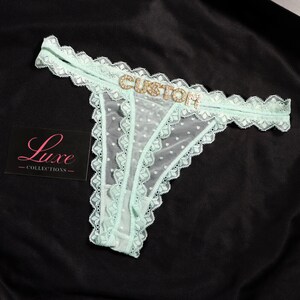 K&ouml;nnte beinhalten: Ein hellminzgr&uuml;ner Tanga aus transparentem Netzstoff mit Punkten, verziert mit einer Spitzenborte an den R&auml;ndern und dem Wort CUSTOM in goldenen Strassbuchstaben am vorderen Bund. Eine schwarze Karte mit dem Text Luxe COLLECTIONS liegt daneben.