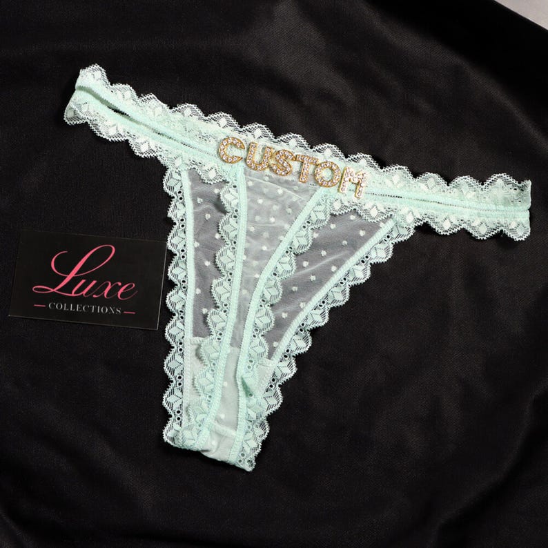 K&ouml;nnte beinhalten: Ein hellminzgr&uuml;ner Tanga aus transparentem Netzstoff mit Punkten, verziert mit einer Spitzenborte an den R&auml;ndern und dem Wort CUSTOM in goldenen Strassbuchstaben am vorderen Bund. Eine schwarze Karte mit dem Text Luxe COLLECTIONS liegt daneben.