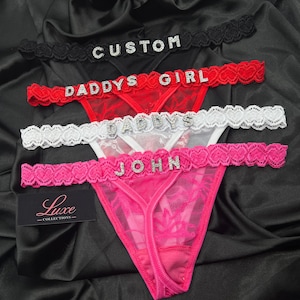 Puede incluir: Cuatro tangas de encaje en negro, rojo, blanco y rosa, cada uno con una cinturilla con patr&oacute;n de corazones y letras de pedrer&iacute;a que deletrean CUSTOM, DADDYS GIRL, DADDYS y JOHN. Incluye una etiqueta rectangular negra con el texto Luxe COLLECTIONS.