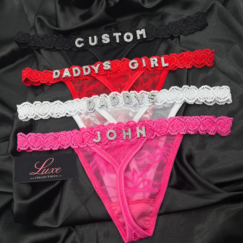 Puede incluir: Cuatro tangas de encaje en negro, rojo, blanco y rosa, cada uno con una cinturilla con patr&oacute;n de corazones y letras de pedrer&iacute;a que deletrean CUSTOM, DADDYS GIRL, DADDYS y JOHN. Incluye una etiqueta rectangular negra con el texto Luxe COLLECTIONS.