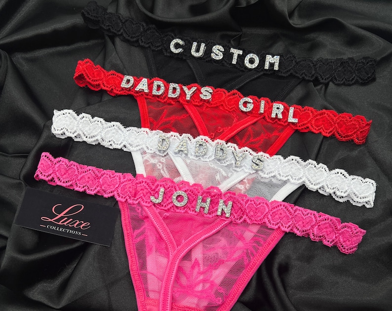 Puede incluir: Cuatro tangas de encaje en negro, rojo, blanco y rosa, cada uno con una cinturilla adornada con pedrer&iacute;a. Las cinturillas muestran el texto CUSTOM, DADDYS GIRL, DADDYS y JOHN en letras plateadas. Una etiqueta negra dice Luxe COLLECTIONS.