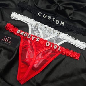 Puede incluir: Tres tangas en negro, blanco y rojo con cinturillas de encaje y letras de pedrer&iacute;a. La tanga negra dice CUSTOM, la blanca DADDYS y la roja DADDYS GIRL. Cada pieza est&aacute; confeccionada con un tejido de malla transparente y delicado.