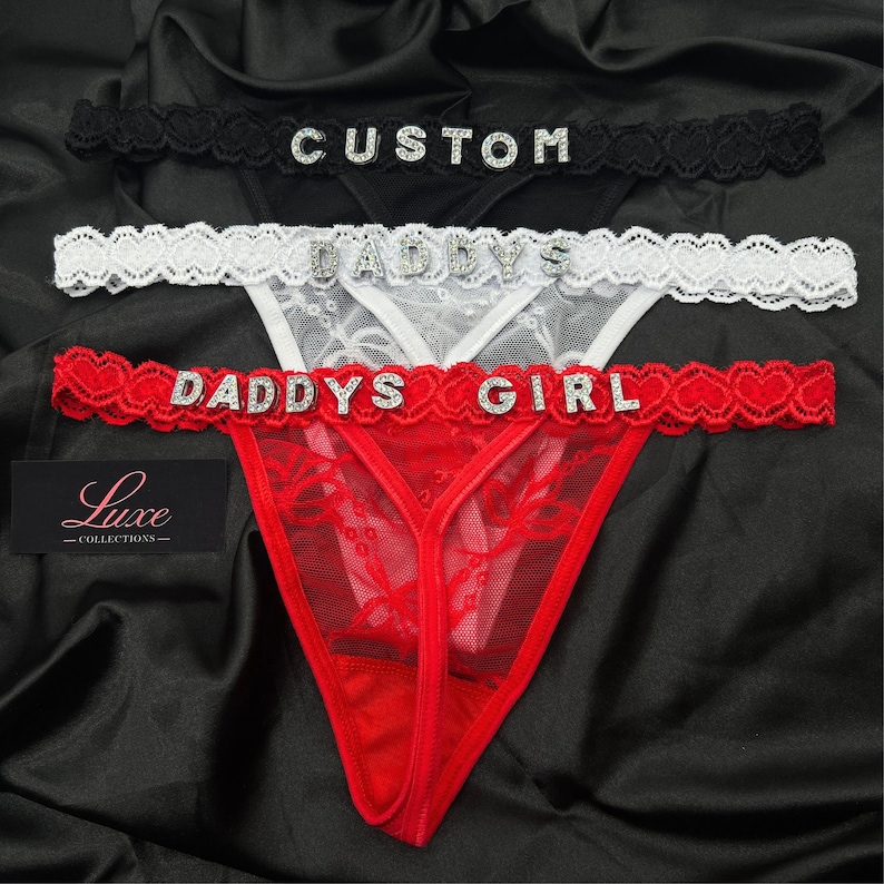 Puede incluir: Tres tangas de encaje en negro, blanco y rojo. Cada uno presenta una cintura festoneada con letras de pedrer&iacute;a. El par negro dice CUSTOM, el blanco dice DADDYS y el rojo dice DADDYS GIRL. Una etiqueta negra muestra el texto Luxe COLLECTIONS.