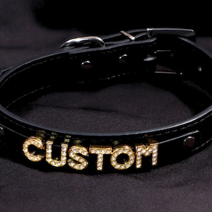 May include: Black leather dog collar with the word "CUSTOM" spelled out in gold-colored, gem-studded letters. The collar has a silver-colored buckle and is secured with rivets. The background is a dark, textured fabric.