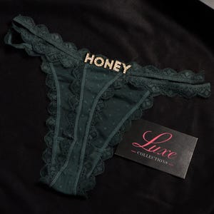 K&ouml;nnte beinhalten: Dunkelgr&uuml;ner Tanga mit Spitzenbesatz und einem zentralen Strass-Schmuckstein, der das Wort HONEY bildet. Eine schwarze rechteckige Karte mit dem rosa und wei&szlig;en Text Luxe COLLECTIONS liegt neben dem Kleidungsst&uuml;ck.