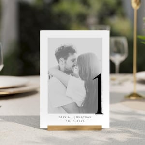 May include: Black and white photo of a couple kissing, with the text "Olivia + Jonathan 10.11.2025" and the number "1" in a large, bold font. This is a table number card for a wedding.