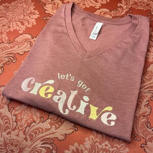 Puede incluir: Una camiseta de cuello en V de color malva con el texto "let's get creative" bordado en amarillo y blanco.