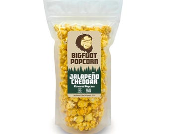 May include: A clear plastic bag filled with yellow popcorn. The bag has a label that says "Bigfoot Popcorn" and "Jalapeño Cheddar Flavored Popcorn".