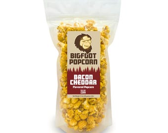 May include: A clear plastic bag filled with yellow popcorn. The bag has a brown label with the text "BIGFOOT POPCORN" and "BACON CHEDDAR Flavored Popcorn" in white text. The label also features a cartoon image of a Bigfoot.