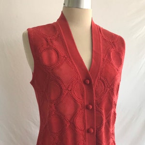May include: A red sleeveless knitted vest with a V-neck and button closure. The vest has a geometric pattern of circles and diamonds.