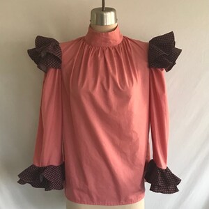 May include: A salmon pink blouse with a high neck and long sleeves. The sleeves and shoulders have black and white patterned ruffles.