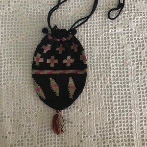 May include: A black crocheted drawstring bag with a pink and beige geometric pattern. The bag has a long black cord and a tassel.