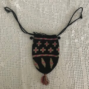 May include: A black crocheted drawstring bag with a pink and white geometric pattern. The bag has a tassel and a long black cord.