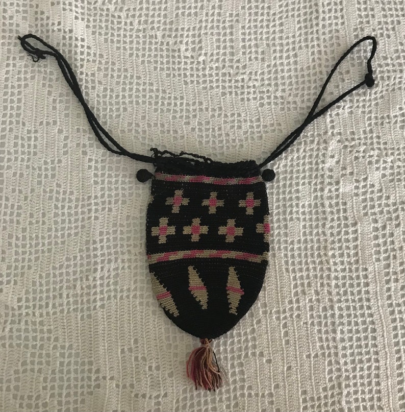 May include: A black crocheted drawstring bag with a pink and white geometric pattern. The bag has a tassel and a long black cord.