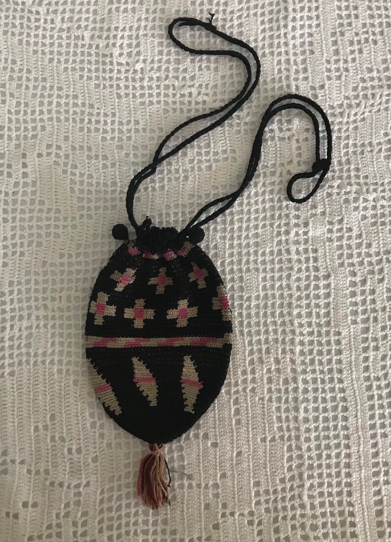 May include: A black crocheted drawstring bag with a pink and beige geometric pattern. The bag has a long black cord and a tassel.