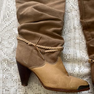 May include: Brown suede knee-high boots with a braided strap detail and a light brown leather toe. The boots have a stacked heel.