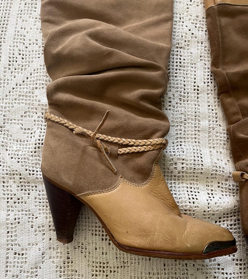 May include: Brown suede knee-high boots with a braided strap detail and a light brown leather toe. The boots have a stacked heel.