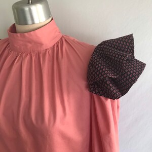 May include: A pink blouse with a high neck and a ruffled, patterned sleeve. The sleeve is made of a dark fabric with a small floral print.