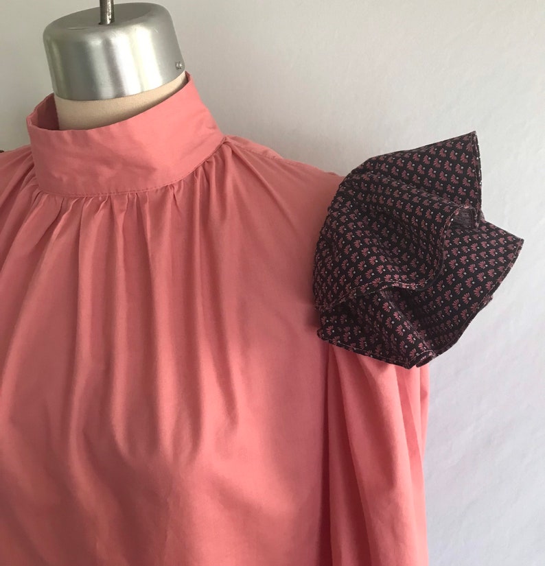 May include: A pink blouse with a high neck and a ruffled, patterned sleeve. The sleeve is made of a dark fabric with a small floral print.