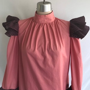 May include: A pink long-sleeved blouse with a high neck and ruffled black and white patterned fabric on the shoulders.