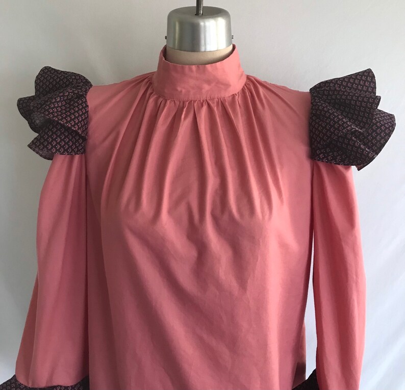 May include: A pink long-sleeved blouse with a high neck and ruffled black and white patterned fabric on the shoulders.