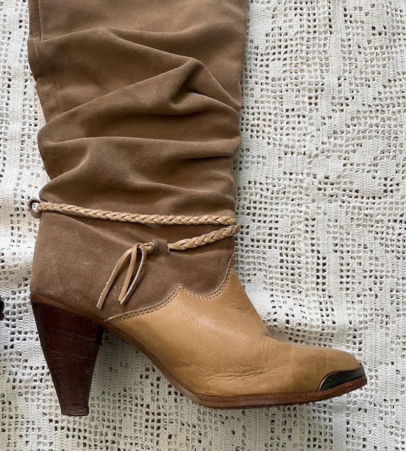 May include: Brown suede and leather knee-high boots with a braided strap and tassel detail. The boots have a stacked heel and a pointed toe.