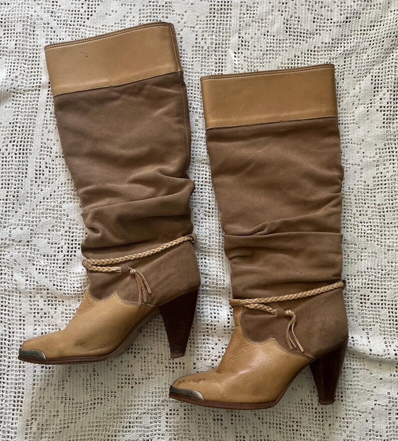 May include: A pair of brown suede knee-high boots with a braided detail around the ankle and a pointed toe.