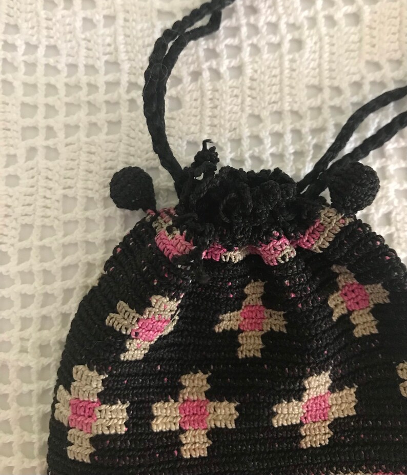 May include: A black crocheted drawstring bag with a pink and beige floral pattern. The bag has a tassel on each side.
