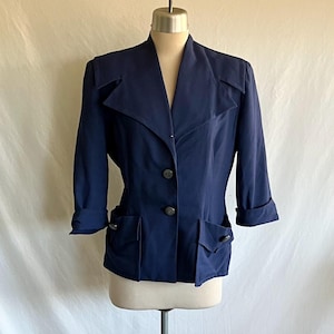 May include: A navy blue blazer with a notched collar, three-quarter sleeves, and two front pockets. The blazer has two dark buttons and is displayed on a mannequin. The sleeves are cuffed.