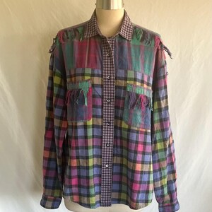 May include: A colorful plaid shirt with a patchwork design. The shirt features a mix of pink, green, purple, and yellow squares. It has fringe detailing on the shoulders and pockets, and a gingham pattern down the center.