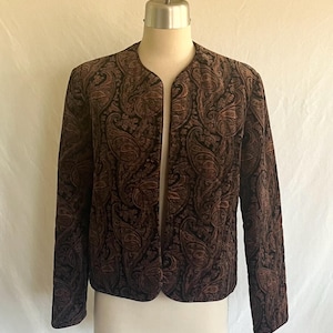 May include: A brown paisley print jacket with a collarless design. The jacket is displayed on a mannequin, showcasing its long sleeves and open front. The fabric has a dark brown and black color scheme.