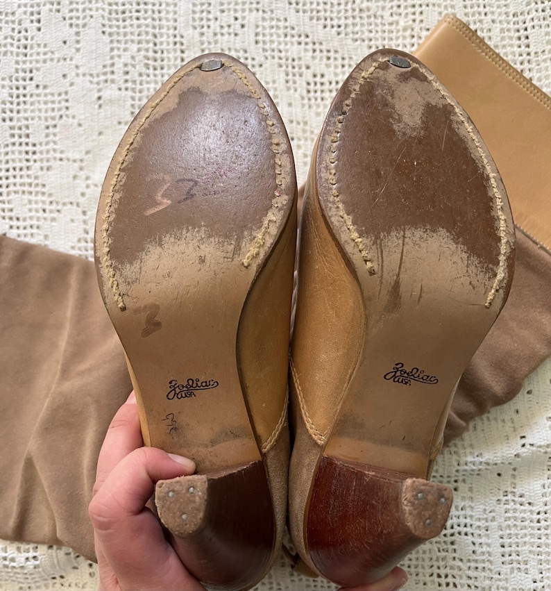 May include: A pair of brown suede knee-high boots with a wooden heel. The boots have a zipper on the inside of the leg and the brand name "Zodiac" is printed on the sole.