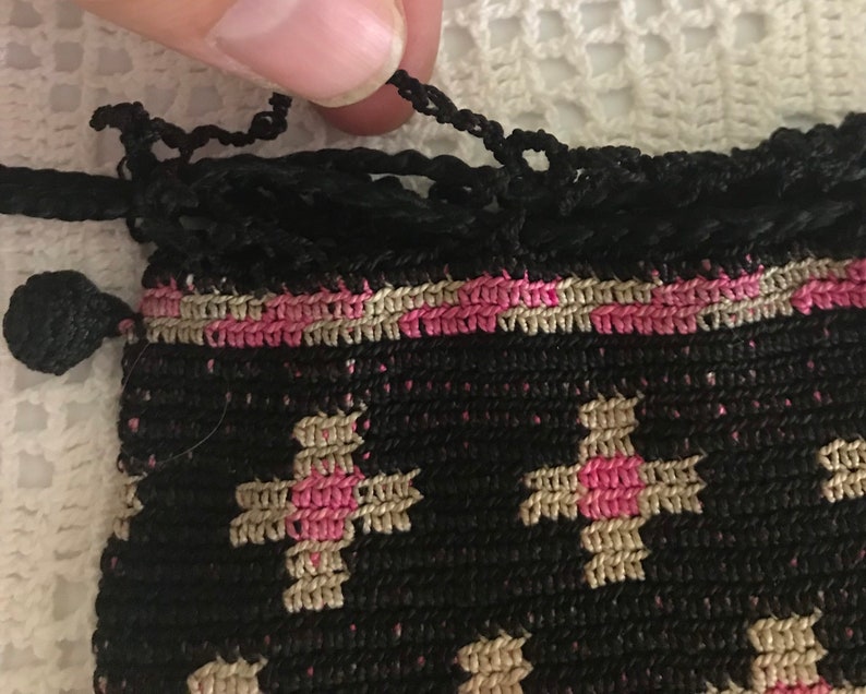 May include: A small black crocheted bag with a pink and beige pattern. The bag has a drawstring closure and a tassel.