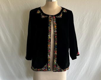 May include: Black velvet jacket with colorful floral embroidery along the collar, front edges, and cuffs. The jacket has three-quarter sleeves and is displayed on a mannequin. The floral embroidery features a variety of colors.