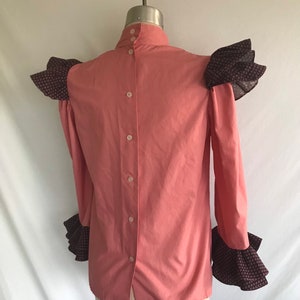 May include: A pink long-sleeved blouse with a high collar and button-up back. The sleeves and shoulders have ruffled trim in a black and white floral print.